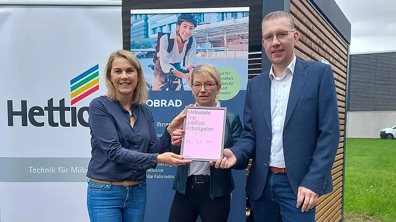 Monika Titze, von der Hettich Gruppe, steht zusammen mit einer Frau und einem Mann vor einem Poster mit dem Schriftzug Hettich und dem Hettich Logo. Sie alle tragen blaue Geschäftskleidung