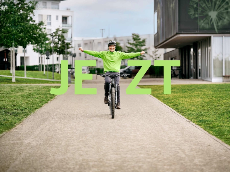 JobRadler fährt strahlend auf seinem Fahrrad mit gestreckten Armen mit Schriftzug "Jetzt"