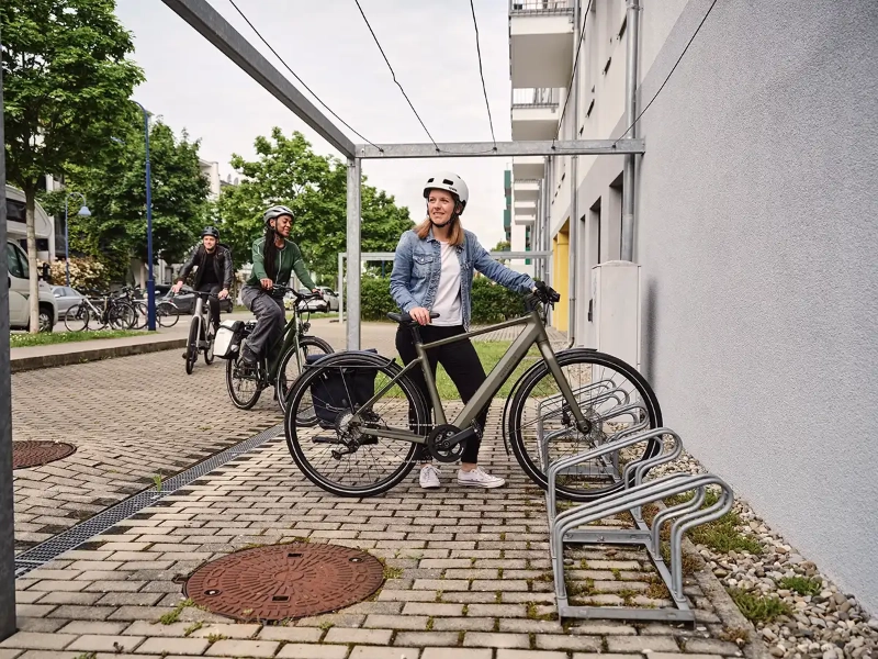 Eine Frau in blauer Jeansjacke parkt ihr grünes E-Bike, eine weitere Frau und ein Mann fahren den Fahrradstellplatz an Eine Frau in blauer Jeansjacke parkt ihr grünes E-Bike, eine weitere Frau und ein Mann fahren den Fahrradstellplatz an